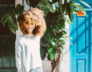 A woman with curly hair smiles in front of a vibrant blue door and lush plant.