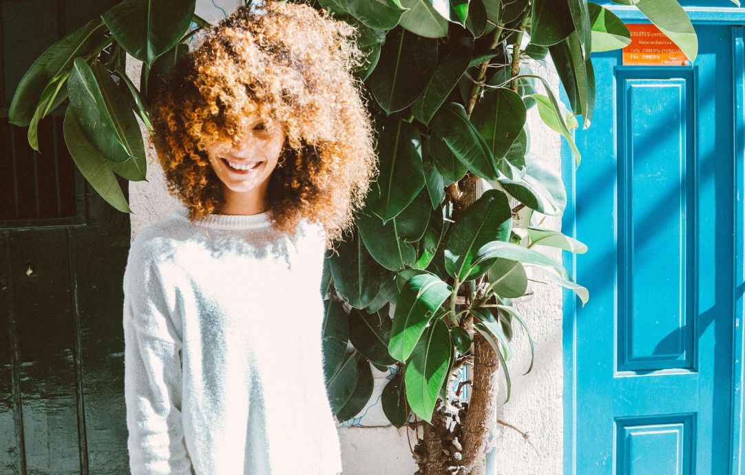 A woman with curly hair smiles in front of a vibrant blue door and lush plant.