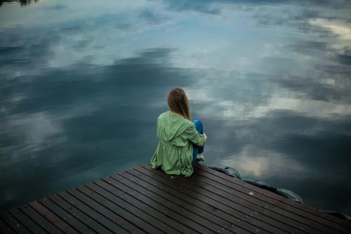 A woman sits on a wooden dock, reflecting by a calm lake under a cloudy sky.